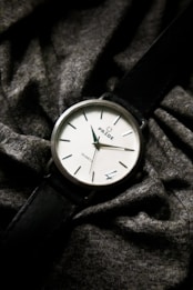 A classic analog wristwatch with a black leather strap and a simple white face displaying the brand name 'PRIDE'. It shows minute and hour hands, with hour markers. Draped across a textured, dark grey fabric which adds a casual and understated elegance to the presentation.
