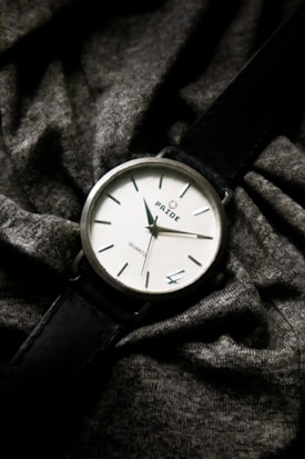 A classic analog wristwatch with a black leather strap and a simple white face displaying the brand name 'PRIDE'. It shows minute and hour hands, with hour markers. Draped across a textured, dark grey fabric which adds a casual and understated elegance to the presentation.