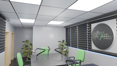 A modern conference room featuring a large table with microphones and green chairs. The room is well-lit with recessed ceiling lights and has dark window blinds. There are potted plants adding greenery, and a sign on the wall reads 'Re-Visitation Mandate' with a microphone graphic.
