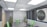 A modern conference room featuring a large table with microphones and green chairs. The room is well-lit with recessed ceiling lights and has dark window blinds. There are potted plants adding greenery, and a sign on the wall reads 'Re-Visitation Mandate' with a microphone graphic.