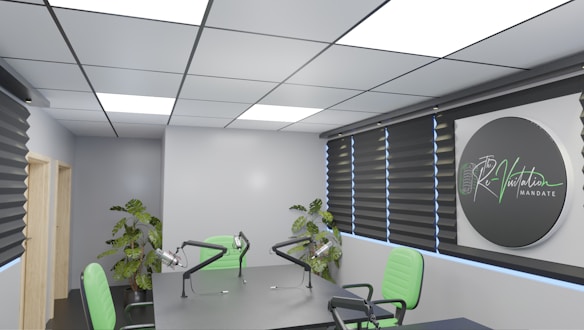 A modern conference room featuring a large table with microphones and green chairs. The room is well-lit with recessed ceiling lights and has dark window blinds. There are potted plants adding greenery, and a sign on the wall reads 'Re-Visitation Mandate' with a microphone graphic.