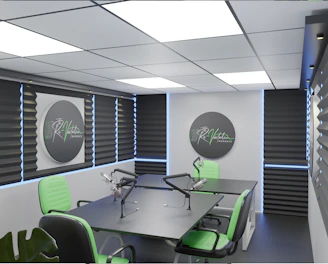 A modern podcast studio with professional microphone setups on a table surrounded by green and black office chairs. The room has acoustic foam on the walls for soundproofing and large square ceiling lights. The walls are predominantly adorned with decorative panels and some branding elements.