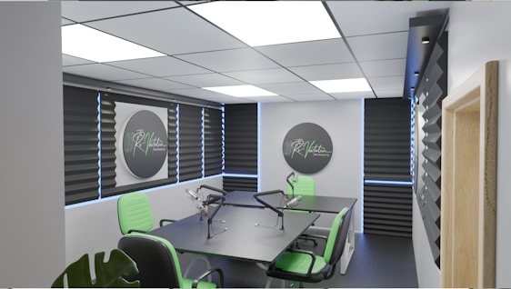 A modern podcast studio with professional microphone setups on a table surrounded by green and black office chairs. The room has acoustic foam on the walls for soundproofing and large square ceiling lights. The walls are predominantly adorned with decorative panels and some branding elements.