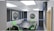 A modern podcast studio with professional microphone setups on a table surrounded by green and black office chairs. The room has acoustic foam on the walls for soundproofing and large square ceiling lights. The walls are predominantly adorned with decorative panels and some branding elements.