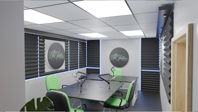 A modern podcast studio with professional microphone setups on a table surrounded by green and black office chairs. The room has acoustic foam on the walls for soundproofing and large square ceiling lights. The walls are predominantly adorned with decorative panels and some branding elements.