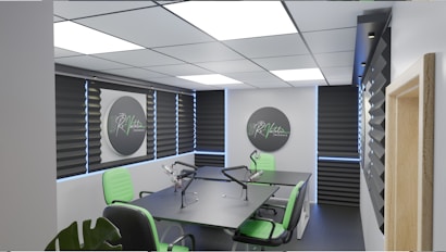 A modern podcast studio with professional microphone setups on a table surrounded by green and black office chairs. The room has acoustic foam on the walls for soundproofing and large square ceiling lights. The walls are predominantly adorned with decorative panels and some branding elements.