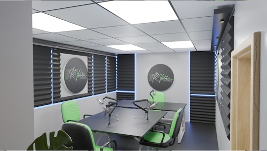 A modern podcast studio with professional microphone setups on a table surrounded by green and black office chairs. The room has acoustic foam on the walls for soundproofing and large square ceiling lights. The walls are predominantly adorned with decorative panels and some branding elements.