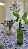 A dining table is set with decorative elements including a green glass bottle with a leafy plant and a potted cactus on a hammered metallic bowl. The tablecloth features a blue and white floral and geometric pattern, and there are neatly arranged glasses and cutlery. A colorful metal wall decoration with bird and leaf motifs is visible on the wall.