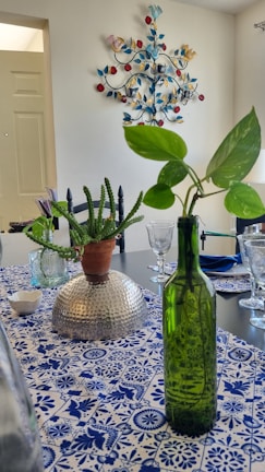 A dining table is set with decorative elements including a green glass bottle with a leafy plant and a potted cactus on a hammered metallic bowl. The tablecloth features a blue and white floral and geometric pattern, and there are neatly arranged glasses and cutlery. A colorful metal wall decoration with bird and leaf motifs is visible on the wall.