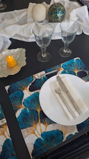 A sophisticated table setting features two clear goblets, a white plate with a silver fork and knife, and white napkin on a patterned placemat with blue and gold leaf designs. Nearby, a decorative display includes a textured white orb and a green mosaic sphere on a white fabric. A yellow candle stands on a glass holder.