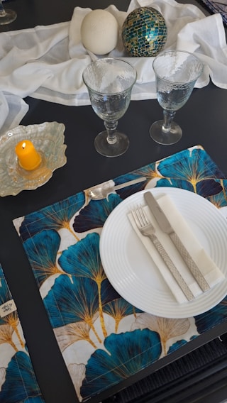 A sophisticated table setting features two clear goblets, a white plate with a silver fork and knife, and white napkin on a patterned placemat with blue and gold leaf designs. Nearby, a decorative display includes a textured white orb and a green mosaic sphere on a white fabric. A yellow candle stands on a glass holder.