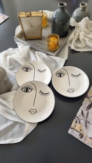 A set of three white plates, each adorned with minimalist black line art depicting abstract faces, is placed on a dark surface. Two yellow glass candle holders and a vintage clock are on a gray tray alongside a gray vase, exuding an elegant and artistic ambiance. White fabric is draped around the objects, adding a soft and delicate touch.