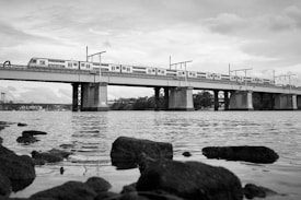 A black and white image of a train traveling across a bridge over a body of water. The bridge is supported by large columns, and in the foreground, rocks are visible in the water. The sky is cloudy, creating a reflective surface on the water.