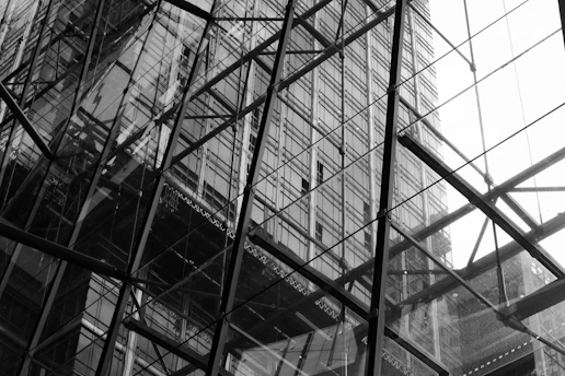 A monochromatic photograph capturing the reflection of a high-rise building on the glass windows of another structure. The intricate network of metal beams and frames create a grid-like pattern, adding a sense of modern architecture and urban environment.
