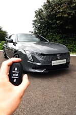 A sleek gray hybrid car, identifiable by the badge and model name on the front, is parked on a road. A hand holding a car key fob is visible in the foreground, indicating an interaction with the vehicle. The background features lush green foliage, providing a contrasting backdrop to the car's modern design.