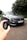 A sleek gray hybrid car, identifiable by the badge and model name on the front, is parked on a road. A hand holding a car key fob is visible in the foreground, indicating an interaction with the vehicle. The background features lush green foliage, providing a contrasting backdrop to the car's modern design.