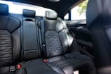 The interior of a modern car displaying the rear passenger seats. The seats are upholstered in black leather with a textured pattern in the center. The car appears to be clean and well-maintained, with the seat belts properly arranged.