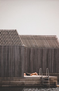A person is sunbathing on a wooden deck next to a structure with a series of uniformly sloped roofs. There is a ladder leading down into water, suggesting this is a waterside or poolside setting.