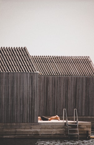 A person is sunbathing on a wooden deck next to a structure with a series of uniformly sloped roofs. There is a ladder leading down into water, suggesting this is a waterside or poolside setting.