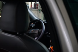 The image captures the interior of a vehicle viewed from the back seat. The focus is on the steering wheel, which has a logo in the center. The dashboard and part of the driver's seat are visible, with subtle reflections and soft lighting creating a calm atmosphere.