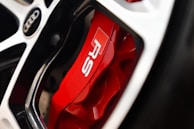 Close-up view of a car wheel featuring a red brake caliper with the letters 'RS' in white. The image also includes part of a multi-spoke alloy wheel and the logo of the car manufacturer.