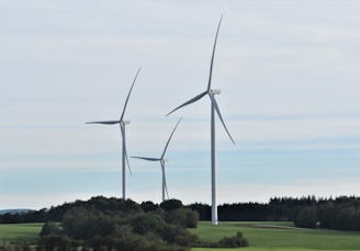 Three large wind turbines stand in a green landscape with a backdrop of trees and an overcast sky. The turbines are evenly spaced and dominate the scene, providing a sense of modern technology amidst nature.