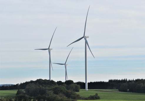 Three large wind turbines stand in a green landscape with a backdrop of trees and an overcast sky. The turbines are evenly spaced and dominate the scene, providing a sense of modern technology amidst nature.