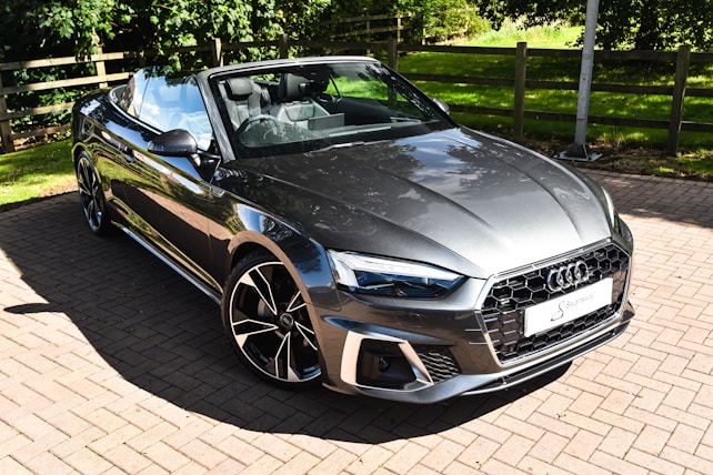 A sleek, modern gray convertible car parked on a sunlit brick driveway. The car features a stylish design with a prominent front grille and sharp headlights. The black and silver alloy wheels complement the metallic body, and the top is down, revealing a luxurious black interior. Lush green trees and a wooden fence are visible in the background, adding a natural touch to the scene.