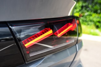 Detail shot of a car's rear LED tail light, featuring sleek and modern red and orange light strips enclosed within a glossy, dark casing. The background includes blurred greenery, indicating the car is outdoors.
