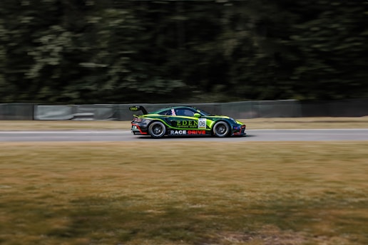 A sports car with a sleek design is racing on a track, surrounded by blurred scenery that implies high speed. The car features vibrant green and black colors with visible sponsorship logos, including a number 06 on its side. It is positioned amidst an environment with green trees and grass.