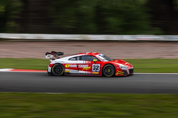 A high-speed sports car races along a track, its vibrant red color and sleek design emphasized by the motion blur that conveys its velocity. The car is branded with various sponsor logos, and the surrounding track and greenery create a scenic backdrop.