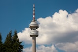 A tall communication tower with a red and white antenna extends into a clear blue sky. Large, fluffy white clouds are visible in the background, with green pine trees at the left edge.
