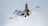 A fighter jet with sleek, modern camouflage patterns flies in the sky. The jet's engines emit fiery, orange exhaust, indicating high speed. The sky is overcast with a uniform gray tone.