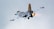A fighter jet with sleek, modern camouflage patterns flies in the sky. The jet's engines emit fiery, orange exhaust, indicating high speed. The sky is overcast with a uniform gray tone.
