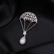 A delicate piece of jewelry shaped like a parachute, adorned with small gemstones and featuring a pear-shaped pearl at the bottom. The jewelry is set against a black, silky fabric background that enhances its luxurious appearance.