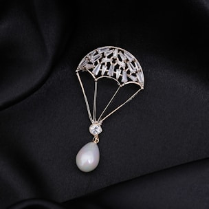 A delicate piece of jewelry shaped like a parachute, adorned with small gemstones and featuring a pear-shaped pearl at the bottom. The jewelry is set against a black, silky fabric background that enhances its luxurious appearance.
