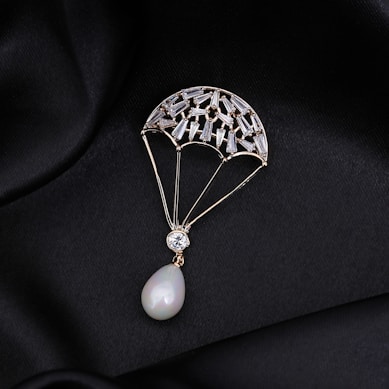 A delicate piece of jewelry shaped like a parachute, adorned with small gemstones and featuring a pear-shaped pearl at the bottom. The jewelry is set against a black, silky fabric background that enhances its luxurious appearance.