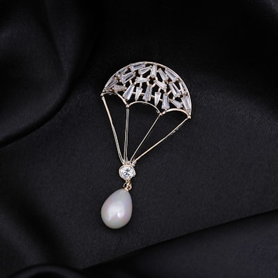 A delicate piece of jewelry shaped like a parachute, adorned with small gemstones and featuring a pear-shaped pearl at the bottom. The jewelry is set against a black, silky fabric background that enhances its luxurious appearance.
