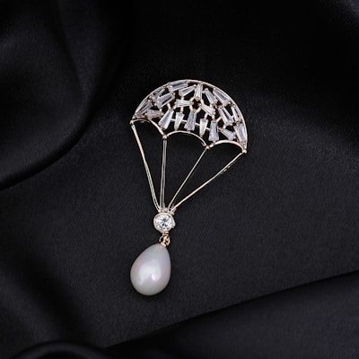 A delicate piece of jewelry shaped like a parachute, adorned with small gemstones and featuring a pear-shaped pearl at the bottom. The jewelry is set against a black, silky fabric background that enhances its luxurious appearance.