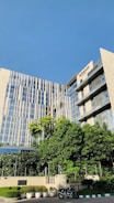 A modern office building with a mix of glass and concrete design. The building has a large, green garden area in the foreground with multiple potted plants and a motorcycle parked on the side. The facade of the building features horizontal and vertical lines reflecting the sunny sky.