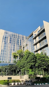 A modern office building with a mix of glass and concrete design. The building has a large, green garden area in the foreground with multiple potted plants and a motorcycle parked on the side. The facade of the building features horizontal and vertical lines reflecting the sunny sky.