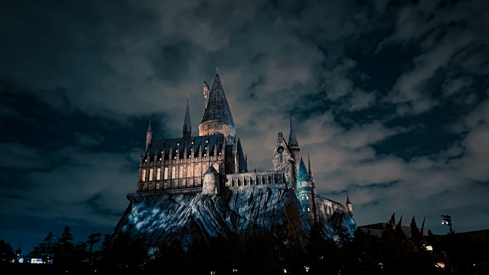 A large, gothic-style castle is perched on a rocky hill under a dramatic, cloud-covered night sky. The castle is illuminated, with its windows glowing warmly, contrasting against the dark, moody atmosphere of the night. Tall spires and intricate architecture add to its imposing and mystical appearance.