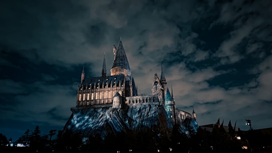 A large, gothic-style castle is perched on a rocky hill under a dramatic, cloud-covered night sky. The castle is illuminated, with its windows glowing warmly, contrasting against the dark, moody atmosphere of the night. Tall spires and intricate architecture add to its imposing and mystical appearance.