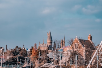 A picturesque scene featuring a castle with tall spires in the background and charming European-style houses in the foreground. Trees with autumn foliage add a warm contrast to the gray and beige tones of the buildings. The sky is clear with a soft blue hue.