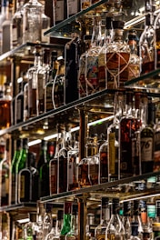A collection of assorted liquor bottles displayed on glass shelves, arranged in a well-lit setting with a variety of shapes, sizes, and labels on the bottles. The shelves are crafted from reflective materials, enhancing the scarcity and selection of the spirits.