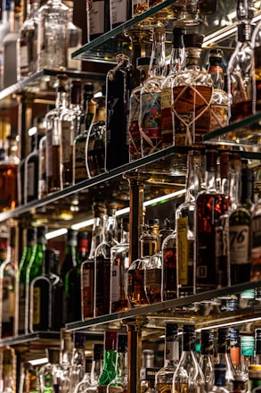 A collection of assorted liquor bottles displayed on glass shelves, arranged in a well-lit setting with a variety of shapes, sizes, and labels on the bottles. The shelves are crafted from reflective materials, enhancing the scarcity and selection of the spirits.