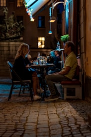 A couple is sitting at a small outdoor caf&eacute; table on a cobblestone street, engaged in conversation. The setting is illuminated by colorful string lights hanging from an awning and the warm glow from nearby windows. Several other patrons can be seen in the background, adding to the lively ambiance of the evening scene.