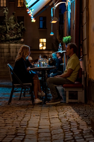 A couple is sitting at a small outdoor caf&eacute; table on a cobblestone street, engaged in conversation. The setting is illuminated by colorful string lights hanging from an awning and the warm glow from nearby windows. Several other patrons can be seen in the background, adding to the lively ambiance of the evening scene.