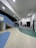 A modern interior space featuring a winding pathway with a multicolored floor in shades of blue, green, and beige. The ceiling is high with recessed lighting and fans. On the left, there is a set of stairs leading up, painted in a light blue color. The walls are adorned with geometric designs, including a red rectangle and various arch shapes in grey tones. A person is seen walking along the pathway.