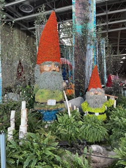 Two large gnome sculptures are adorned with colorful plants and flowers. The gnomes have long grey beards and bright orange hats, surrounded by lush greenery and ferns. The setting appears to be an indoor space with natural elements and decorative pillars in the background.